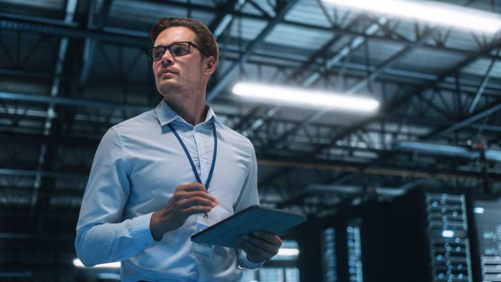 Handsome IT Specialist Using Tablet Computer in Data Center, Looking at the Screen. Succesful Businessman and e-Business Entrepreneur Overlooking Server Farm Cloud Computing Facility.