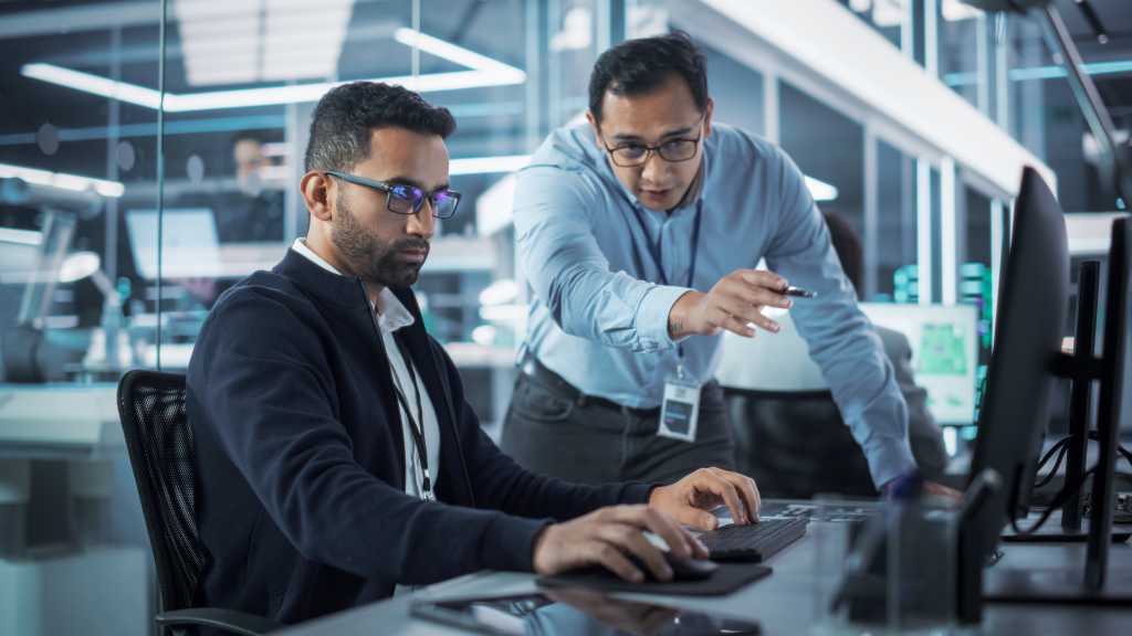 Male Project Supervisor Talks to an Industry 4 Engineer Who Works on Desktop Computer in Robotics Startup Industrial Office. Software Developer Discussing Work Related Tasks During a Meeting.