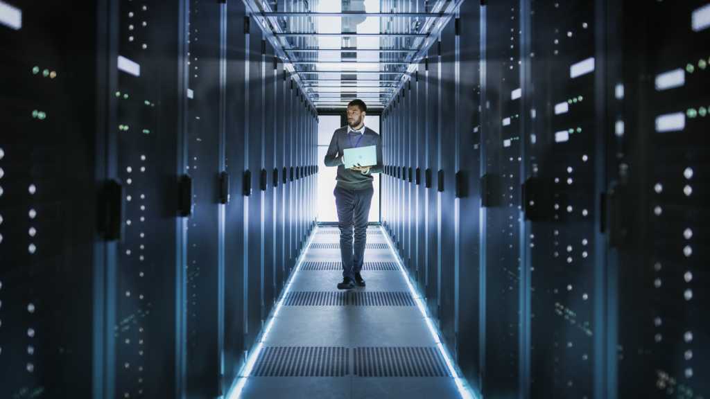 IT Technician Works on Laptop in Big Data Center full of Rack Servers. He Runs Diagnostics and Maintenance, Sets System Up.